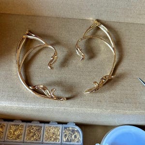 May include: Two gold ear cuffs with a delicate, intricate design. The cuffs are made of metal and have a curved shape.