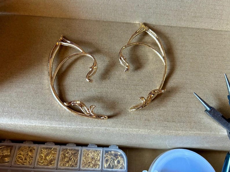 May include: Two gold ear cuffs with a delicate, intricate design. The cuffs are made of metal and have a curved shape.
