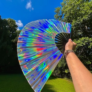 May include: A large handheld folding fan featuring a reflective, iridescent material that displays a spectrum of rainbow colors. The fan is constructed with a black frame and ribs, creating a stark contrast against the shimmering, metallic surface.