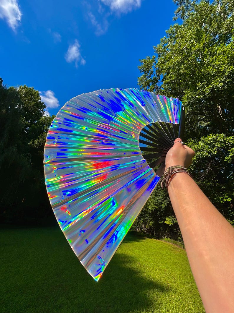May include: A large handheld folding fan featuring a reflective, iridescent material that displays a spectrum of rainbow colors. The fan is constructed with a black frame and ribs, creating a stark contrast against the shimmering, metallic surface.