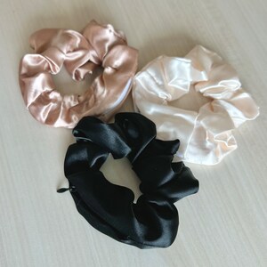 May include: Three satin scrunchies in the image. One is black, one is a light beige, and the other is a pale pink. The black scrunchie has a zipper. These hair accessories are designed to hold hair in place.