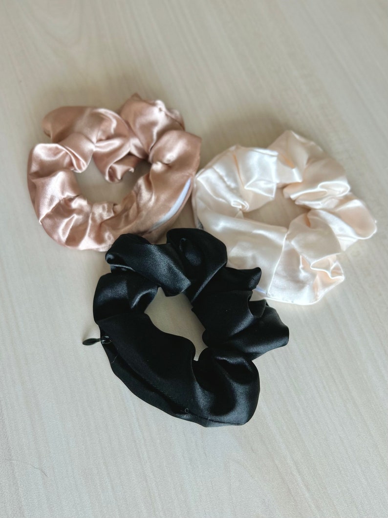 May include: Three satin scrunchies in the image. One is black, one is a light beige, and the other is a pale pink. The black scrunchie has a zipper. These hair accessories are designed to hold hair in place.