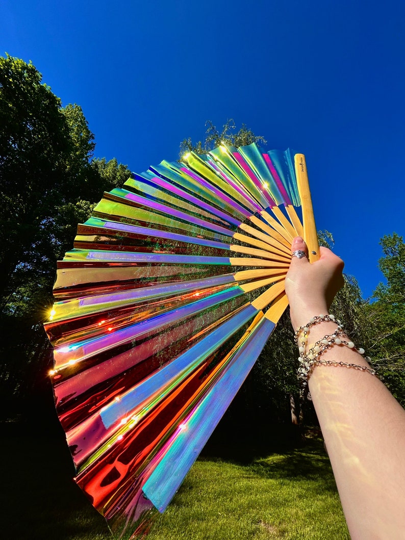 May include: A large hand fan featuring iridescent, holographic material that reflects a spectrum of colors including red, orange, yellow, green, and blue. The fan has a light-colored bamboo frame and includes a small logo printed on the handle.