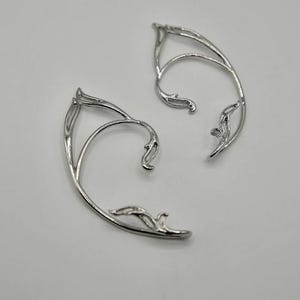 May include: A pair of silver ear cuffs with a delicate, leafy design. The cuffs are curved to fit the shape of the ear.
