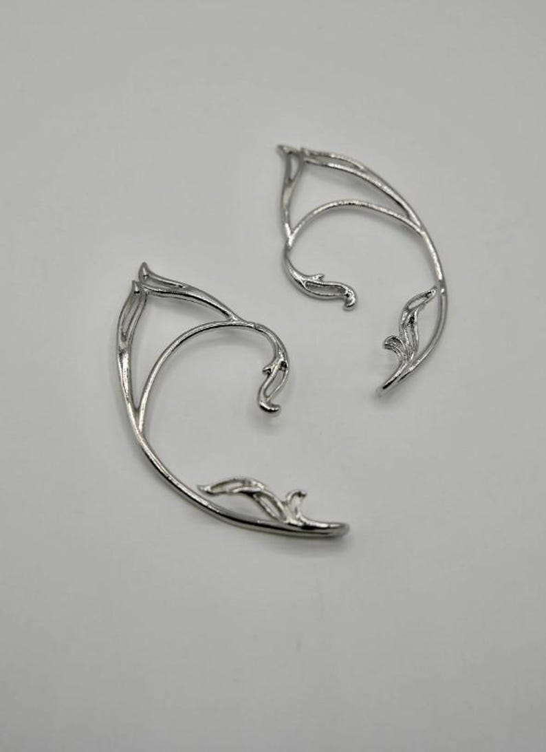May include: A pair of silver ear cuffs with a delicate, leafy design. The cuffs are curved to fit the shape of the ear.