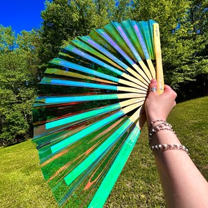 May include: A handheld folding fan featuring a translucent, iridescent material that shifts between shades of green, blue, and purple. The fan is constructed with light-colored wooden ribs and includes a small logo printed on the handle.