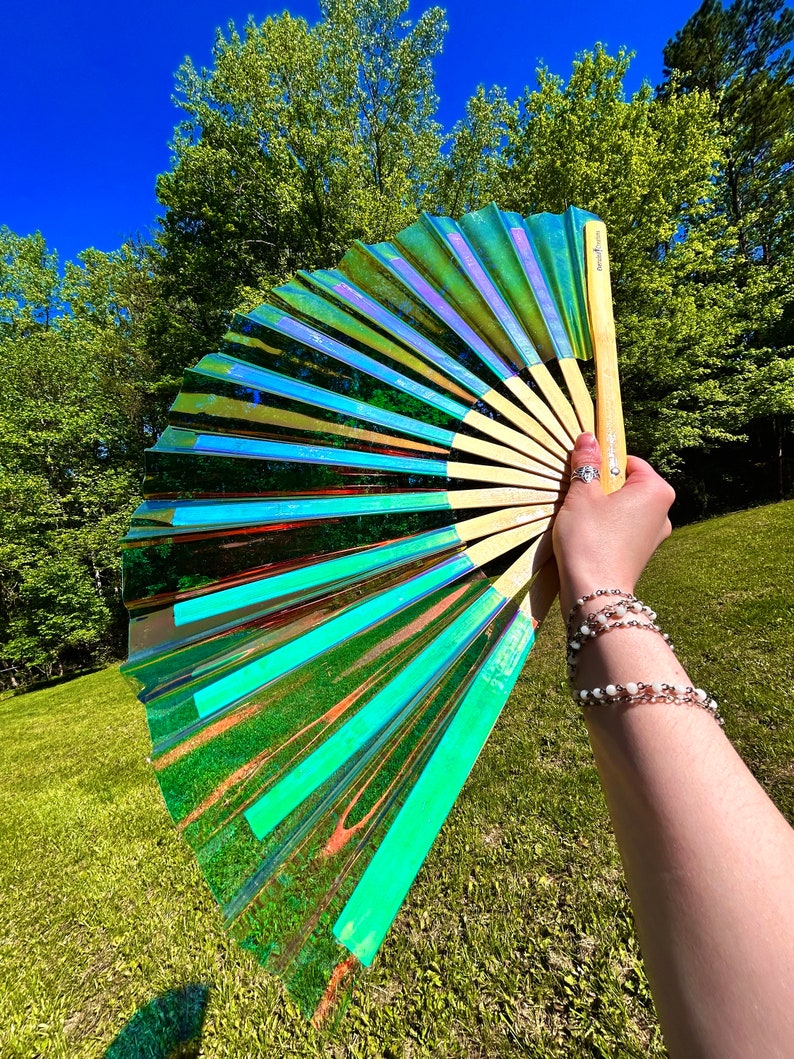 May include: A handheld folding fan featuring a translucent, iridescent material that shifts between shades of green, blue, and purple. The fan is constructed with light-colored wooden ribs and includes a small logo printed on the handle.