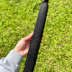 May include: A long, narrow, black fabric sleeve featuring a repeating geometric Greek key pattern. The cylindrical case is constructed from a textured material and includes a black drawstring closure at the top to secure the contents inside.