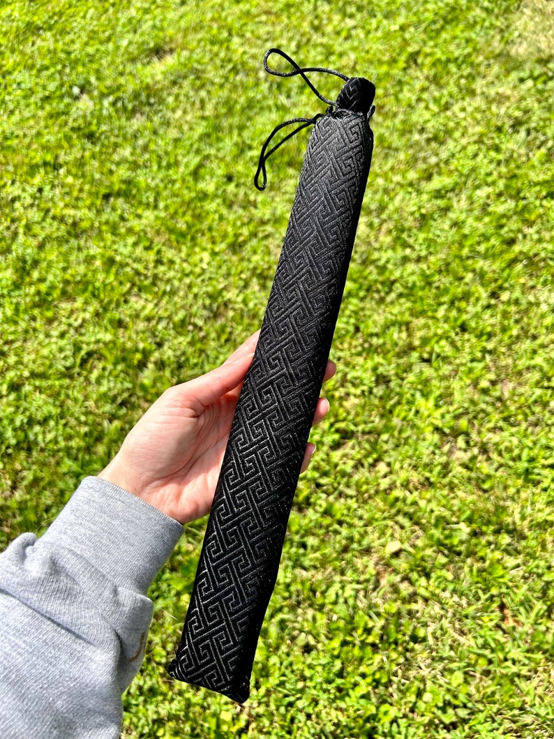 May include: A long, narrow, black fabric sleeve featuring a repeating geometric Greek key pattern. The cylindrical case is constructed from a textured material and includes a black drawstring closure at the top to secure the contents inside.