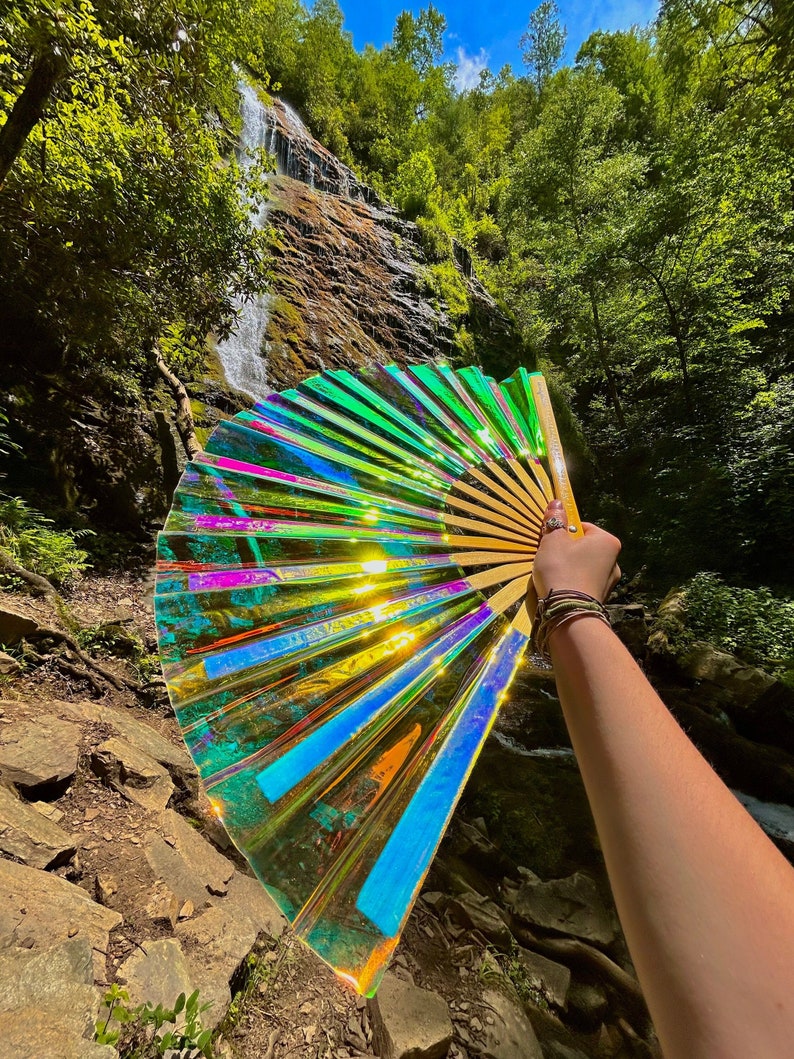 May include: A large handheld folding fan featuring a translucent, iridescent material that shifts between vibrant shades of green, blue, and gold. The fan is constructed with a series of light-colored wooden ribs that radiate from a central pivot point.