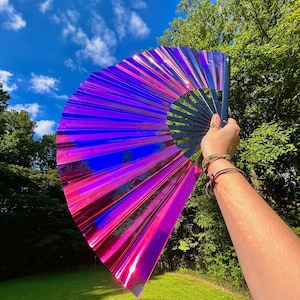 May include: A large handheld folding fan featuring a vibrant, iridescent material that shifts between shades of magenta, purple, and blue. The fan is constructed with a black frame and pleated, reflective panels that create a striking light effect.