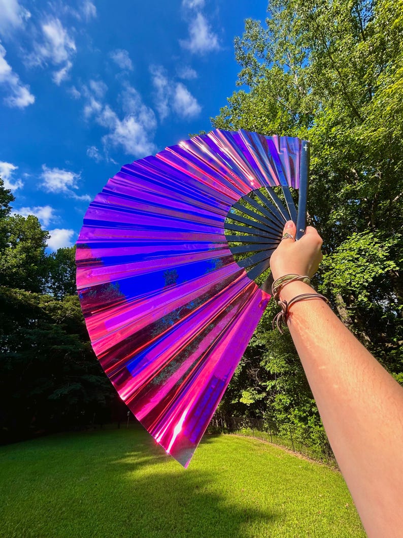 May include: A large handheld folding fan featuring a vibrant, iridescent material that shifts between shades of magenta, purple, and blue. The fan is constructed with a black frame and pleated, reflective panels that create a striking light effect.