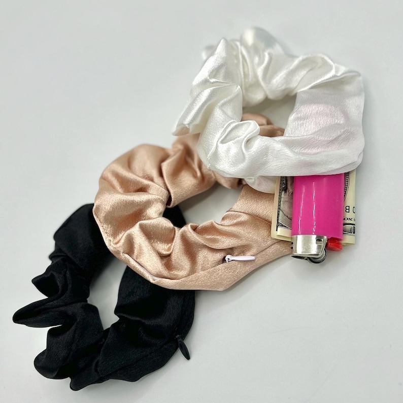 May include: A collection of satin scrunchies in black, blush, and white. One scrunchie has a hidden zipper pocket, and another holds a pink lighter and folded bills. These hair accessories offer a stylish and practical design.