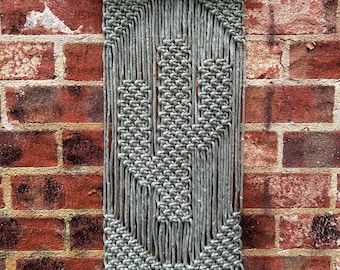May include: A gray macrame wall hanging with a geometric design. The hanging has a wooden dowel at the top and fringe at the bottom.