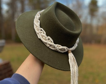 May include: A green felt fedora hat with a white macrame band and tassel. The hat is held in a hand with a blue sleeve.