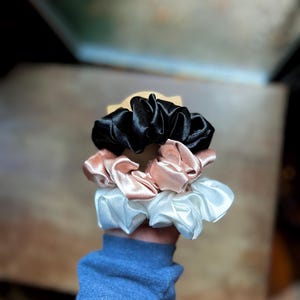 May include: A stack of three satin scrunchies in black, blush pink, and white. The scrunchies are made of a shiny, gathered fabric and are held in a hand. The background is blurred, with a wooden surface and a window visible.