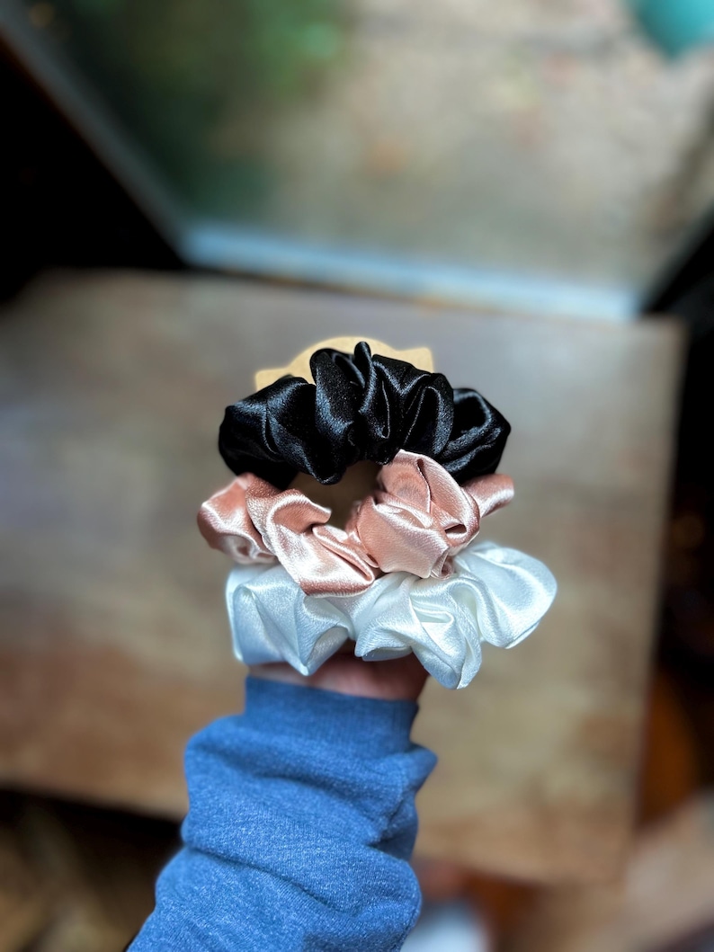 May include: A stack of three satin scrunchies in black, blush pink, and white. The scrunchies are made of a shiny, gathered fabric and are held in a hand. The background is blurred, with a wooden surface and a window visible.