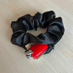 May include: Black satin scrunchie with a hidden zipper pocket. The pocket contains a red lighter, cash, and a small card. The scrunchie is a hair accessory.