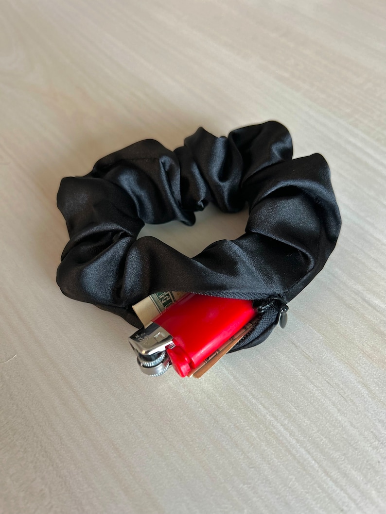 May include: Black satin scrunchie with a hidden zipper pocket. The pocket contains a red lighter, cash, and a small card. The scrunchie is a hair accessory.