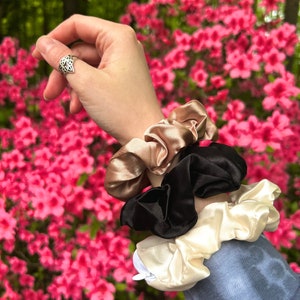 May include: A collection of three satin scrunchies in beige, black, and cream colors. The scrunchies are stacked on top of each other, with a hand wearing a silver ring in the background. The background features pink flowers.