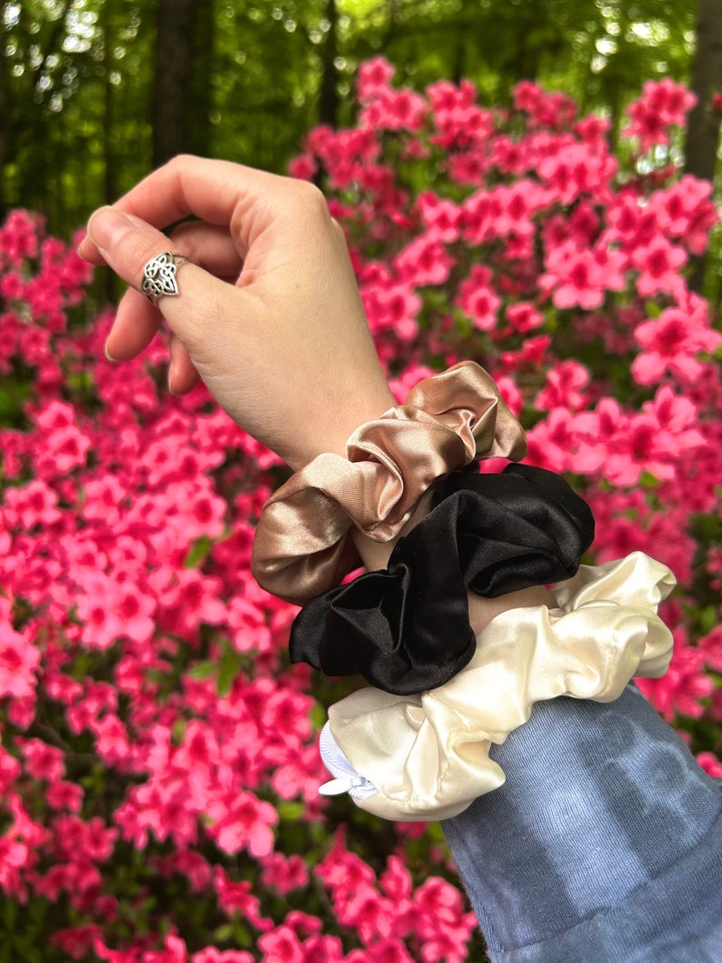 May include: A collection of three satin scrunchies in beige, black, and cream colors. The scrunchies are stacked on top of each other, with a hand wearing a silver ring in the background. The background features pink flowers.
