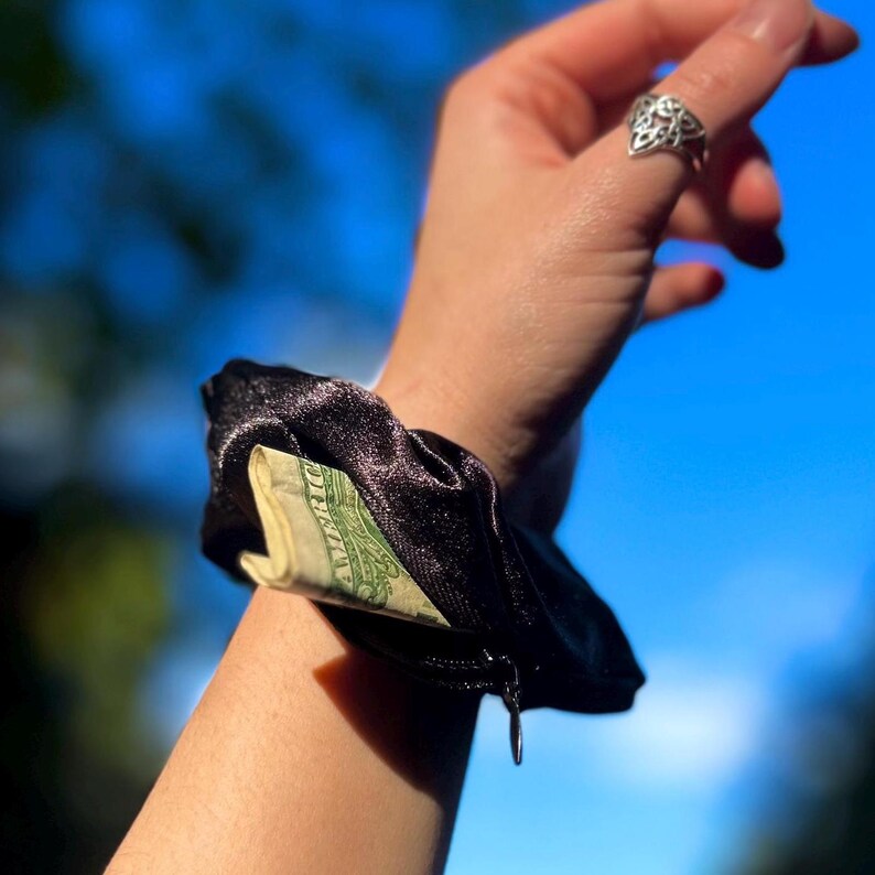 May include: A black satin scrunchie with a hidden zippered pocket, revealing a portion of a dollar bill. The scrunchie is worn on a wrist against a blurred blue sky background.