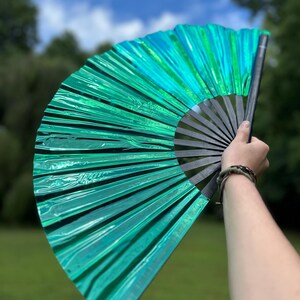 May include: A handheld folding fan featuring a vibrant, metallic emerald green pleated fabric surface. The fan is constructed with a series of black, rigid ribs that radiate from a central pivot point, allowing the fan to open into a wide semicircle.