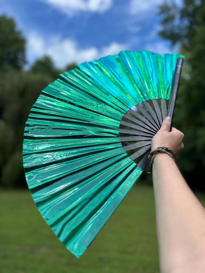 May include: A handheld folding fan featuring a vibrant, metallic emerald green pleated fabric surface. The fan is constructed with a series of black, rigid ribs that radiate from a central pivot point, allowing the fan to open into a wide semicircle.