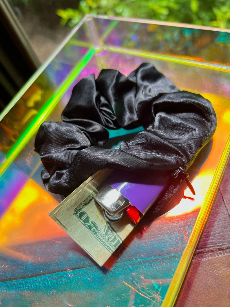 May include: A black satin scrunchie with a twenty-dollar bill and a purple lighter. The scrunchie is made of a shiny fabric and is resting on a reflective surface with a rainbow sheen.