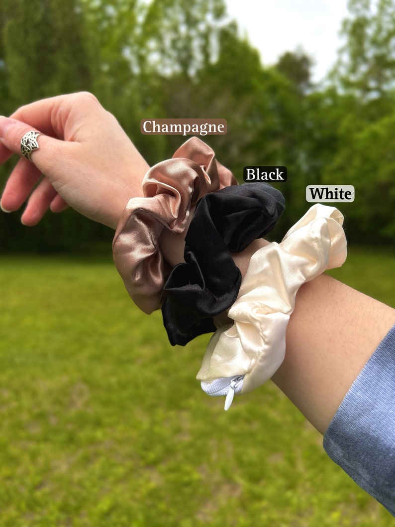 May include: Three satin scrunchies in champagne, black, and white. The scrunchies are displayed on a wrist. The champagne scrunchie is a light, warm beige color. The black scrunchie is a deep, solid black. The white scrunchie is a soft, creamy white.