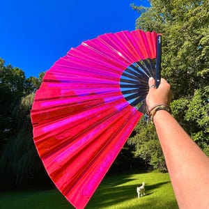 May include: A large, handheld folding fan featuring a vibrant, reflective hot pink material that catches the light. The fan is constructed with black ribs and a black handle, creating a sharp contrast against the bright, metallic pink surface.