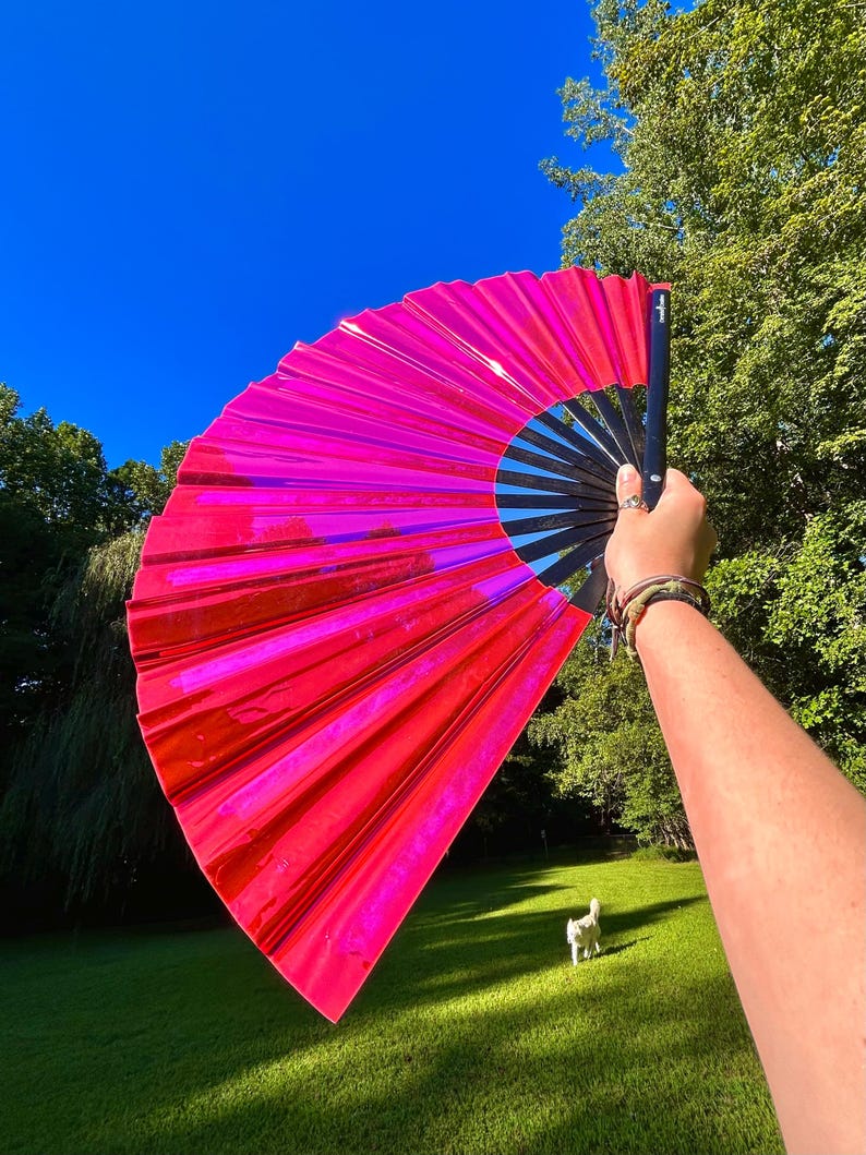 May include: A large, handheld folding fan featuring a vibrant, reflective hot pink material that catches the light. The fan is constructed with black ribs and a black handle, creating a sharp contrast against the bright, metallic pink surface.