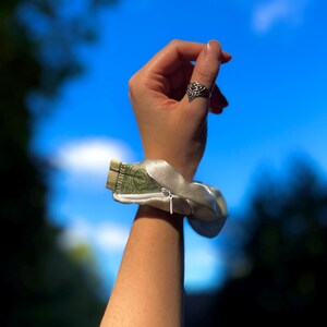 May include: A silver satin wrist wallet with a zipper, holding a folded one-dollar bill. The wristlet is worn on a person's arm, with a silver ring on the index finger. The background is a bright blue sky.