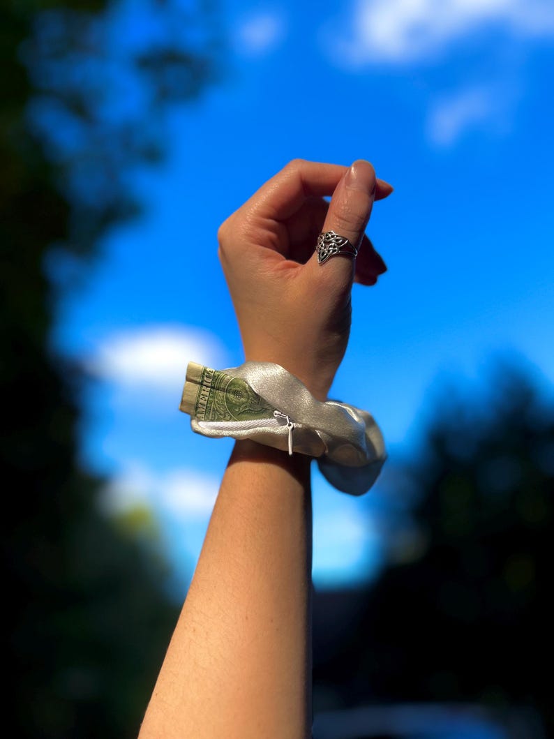 May include: A silver satin wrist wallet with a zipper, holding a folded one-dollar bill. The wristlet is worn on a person's arm, with a silver ring on the index finger. The background is a bright blue sky.