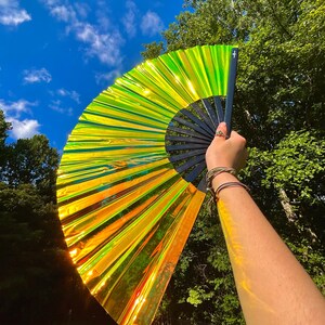 May include: A large, handheld folding fan featuring a vibrant, iridescent lime green and orange metallic finish. The fan is constructed with a black frame and ribs, creating a striking contrast against the reflective, light-catching surface.