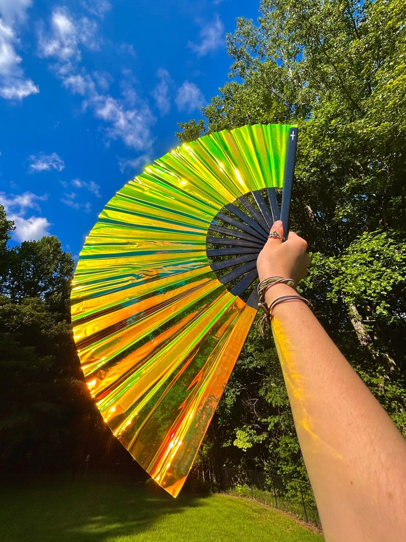 May include: A large, handheld folding fan featuring a vibrant, iridescent lime green and orange metallic finish. The fan is constructed with a black frame and ribs, creating a striking contrast against the reflective, light-catching surface.