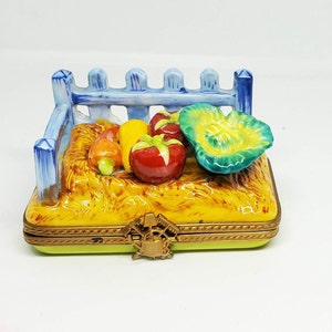 May include: A porcelain trinket box with a blue picket fence design. The box is filled with a hay-like material and features a variety of colorful vegetables, including tomatoes, carrots, and a green cabbage.