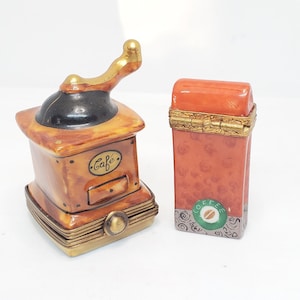 May include: Two small porcelain trinket boxes. One is shaped like a coffee grinder with a gold handle and the word "Cafe" on the front. The other is a rectangular box with a red and orange design and the word "Coffee" on the front.