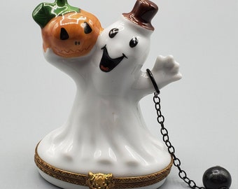 May include: A white ceramic ghost figurine with a brown top hat and a carved orange pumpkin. The ghost has a black chain with a black ball attached. The base has a gold trim and a gold lion head.