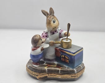 Puede incluir: Figura de cerámica con un conejo y un erizo. El conejo, con un delantal, revuelve una olla en una estufa azul. El erizo observa, sosteniendo un plato. La base es una caja decorativa con bisagras.
