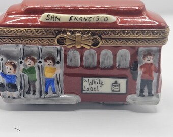 May include: A red and white porcelain trinket box shaped like a San Francisco cable car with a gold trim. The box has a hinged lid with a gold bow. The side of the box has a painted scene of people getting on and off the cable car. The box has the words "San Francisco" and "White Label" painted on the side.