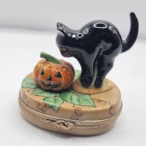 May include: A ceramic Halloween-themed trinket box featuring a black cat and a jack-o'-lantern. The cat is arched, and the pumpkin has a carved face. The box is oval-shaped with a gold-colored trim.