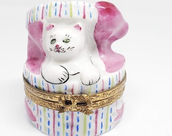 May include: A white porcelain trinket box with a white cat peeking out of a pink and blue striped gift box. The box has a gold hinged lid.