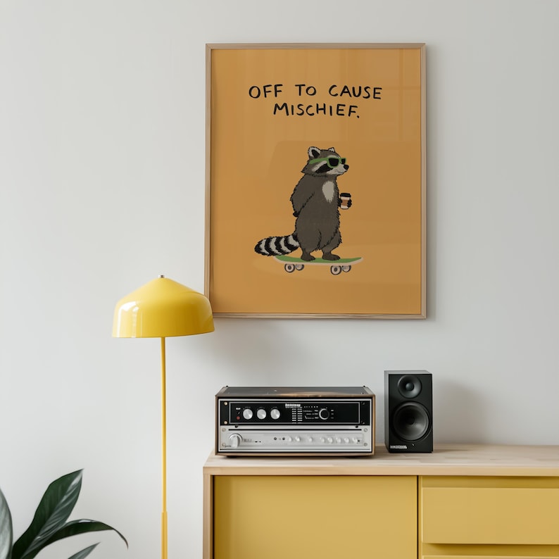 Puede incluir: Una impresi&oacute;n art&iacute;stica enmarcada con un mapache de dibujos animados en una patineta, con gafas de sol y sosteniendo una taza de caf&eacute;. La impresi&oacute;n tiene el texto "OFF TO CAUSE MISCHIEF" sobre un fondo amarillo mostaza. La impresi&oacute;n se muestra encima de un est&eacute;reo y un altavoz.