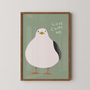 May include: A framed wall art print featuring a stylized illustration of a round white seagull with a dark gray wing and orange feet against a sage green background. The text "LOVE CHIPS ME" is written in white capital letters to the right.