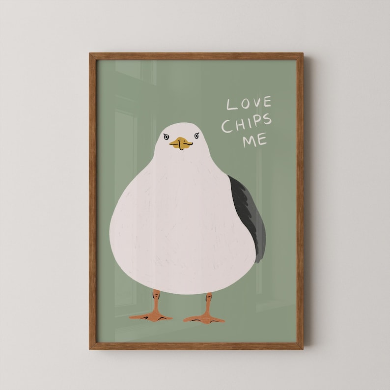May include: A framed wall art print featuring a stylized illustration of a round white seagull with a dark gray wing and orange feet against a sage green background. The text "LOVE CHIPS ME" is written in white capital letters to the right.