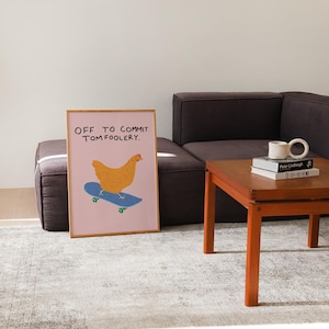 May include: A framed art print featuring a yellow chicken riding a blue skateboard on a pink background. The text "OFF TO COMMIT TOM FOOLERY" is above the chicken. The print is displayed in a room with a brown sofa, a wooden table, and a patterned rug.