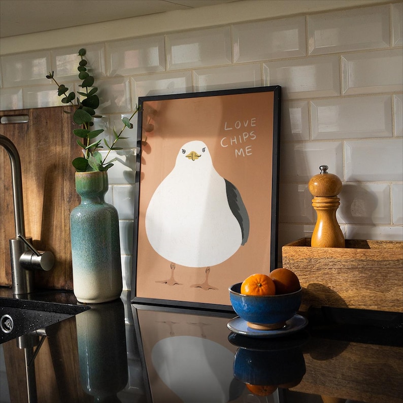 May include: A framed rectangular print featuring a white seagull with a grey wing on a tan background. The text "LOVE CHIPS ME" is written in white in the upper right corner. The print is displayed in a thin black frame on a kitchen counter.