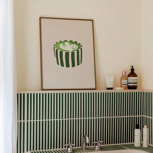 May include: A framed print of a green matcha latte with a brown frame is displayed above a bathtub. The bathroom features green and white striped tile and various toiletries on a shelf.