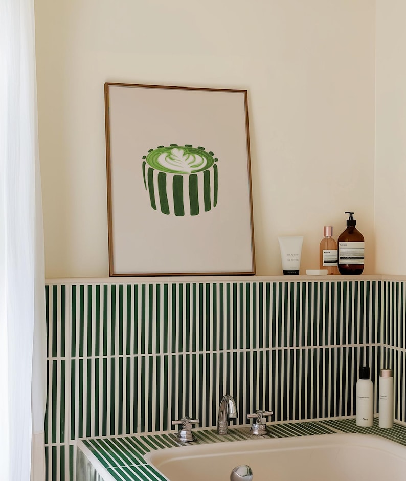 May include: A framed print of a green matcha latte with a brown frame is displayed above a bathtub. The bathroom features green and white striped tile and various toiletries on a shelf.